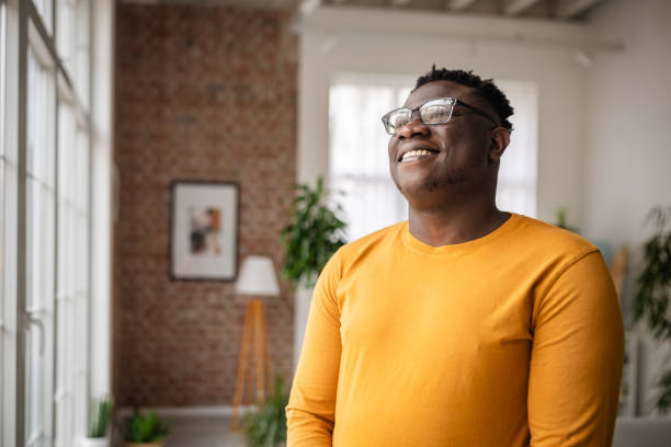 A man wearing a yellow sweater stands smiling in front of a large window, embracing the morning light, inside a cozy home with plants and modern decor elements.