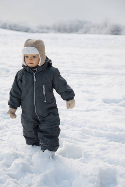 A warmly dressed young child smiling and exploring outdoors in the snow during winter. The child is enjoying the cold weather while wearing a dark snowsuit, gloves, and a hat with ear protection.