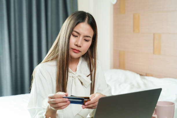 istock Asian female office worker using credit card for online shopping at home, sitting in cozy bedroom with laptop, highlighting convenience, modern technology, and relaxed lifestyle. 2193887607