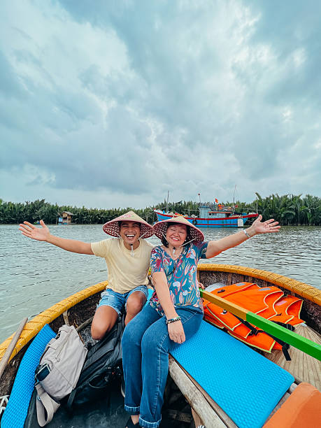 Joyful couple on a boat, embracing the scenic view of the river. Smiling man and woman with Asian conical hats, with paddles and vibrant life jackets aboard.