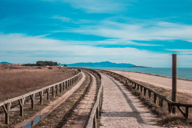 A paved pathway with wooden railings runs along calm coastal waters, bordered by dry grasslands and low mountains under a blue sky with clouds.