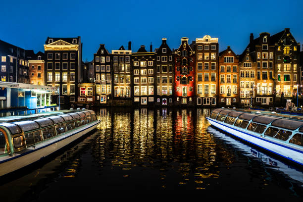 Amsterdam, The Netherlands. January 14th, 2025: A stunning view of Amsterdam's iconic canal houses illuminated at night. The reflection of the warm lights shimmers in the calm water of the canal, flanked by two classic canal cruise boats. The deep blue sky enhances the magical atmosphere of this urban landscape.