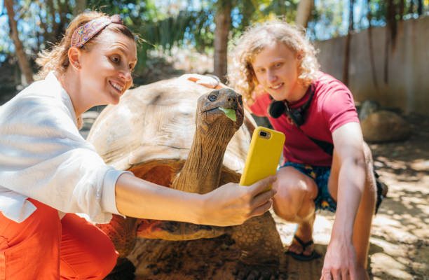 Smiling tourists boy with mom making selfie using cell phone with Aldabra giant tortoise endemic species - one of the largest tortoises in the world in the zoo nature park on Mauritius island.