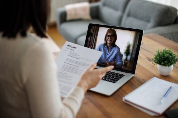 woman attending a virtual job interview while holding her resume - procurar emprego imagens e fotografias de stock