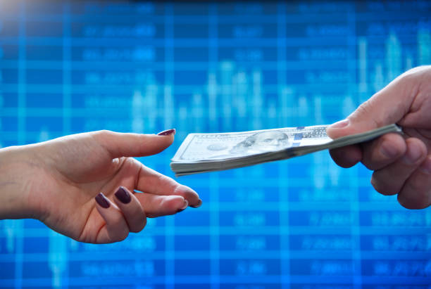 Hand extends a large wad of american dollars to another hand against the background of business charts on the screen