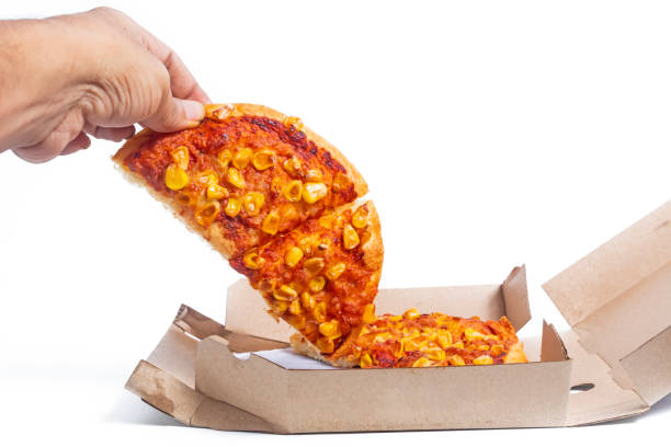 Hand holding a slice of corn topped pizza in cardboard box on white background