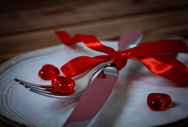 Red glass heart on a fork with a knife wrapped with a red ribbon on a white plate on a brown wooden table, flat lay close-up. Valentine's day concept, festive table, festive table setting.