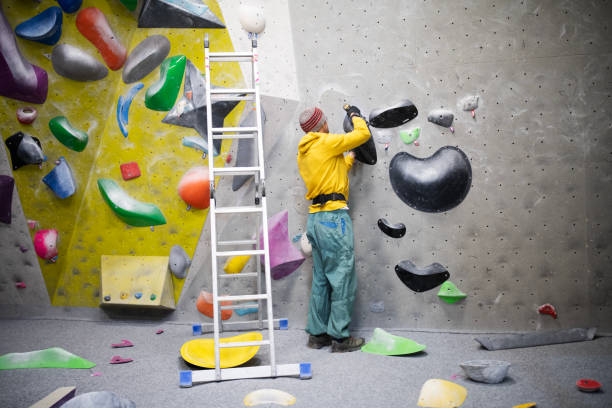 A man is setting up climbing grips on a recently built climbing wall, using a drill to attach them firmly.