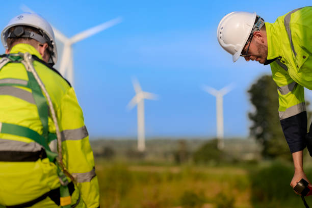 A male engineer in a checkered shirt, orange safety vest, and white helmet stands confidently on a hill at a wind power plant, with modern wind turbines in the background. The clear blue sky and vibrant greenery underline the commitment to sustainable energy solutions. The image symbolizes innovation, clean technology, and the critical role of professionals in advancing the renewable energy sector. Artificial intelligence and digital monitoring systems are becoming essential tools in managing such large-scale operations efficiently and safely