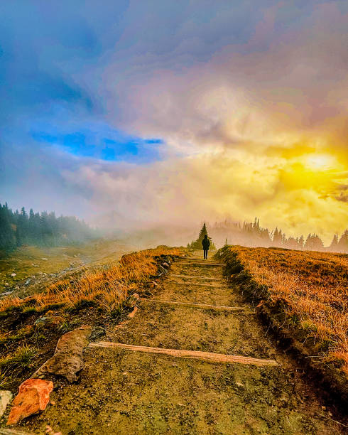 A breathtaking view of a solitary figure walking along a rustic mountain path as the golden sun breaks through the morning mist. The sky is ablaze with hues of yellow, orange, and blue, creating a striking contrast with the rugged trail and surrounding wilderness. This awe-inspiring scene captures the beauty of a quiet journey in nature, symbolizing peace, adventure, and reflection.