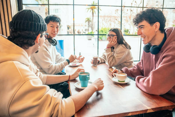 Four young multiracial students are sitting at a table in a coffee shop, drinking coffee and chatting happily together, enjoying their free time and relaxing after a day at university
