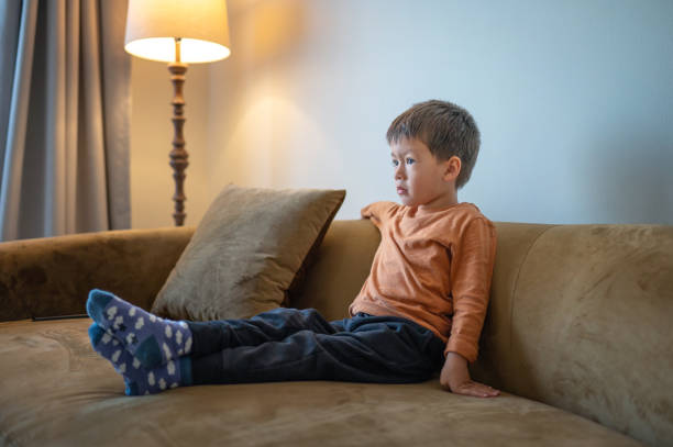 istock Child with orange shirt sitting on brown sofa facing forward a screen  alone. 2193987227