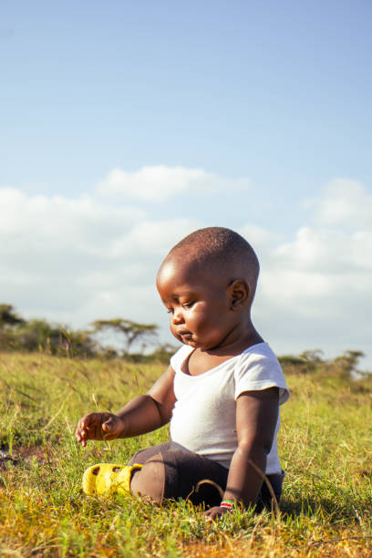 East African child in camouflage print attire sits thoughtfully against yellow fabric backdrop in rural Kenyan location, wearing casual everyday clothing.