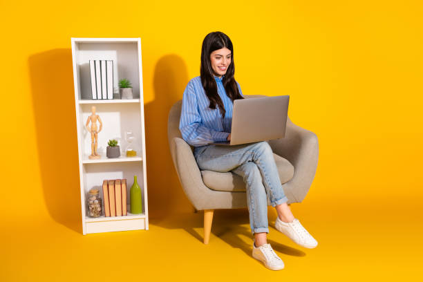istock Young woman using laptop on modern chair after home study with bookshelf on bright yellow background 2193990021