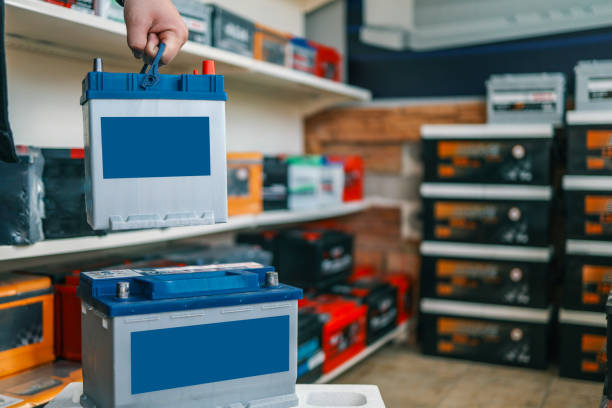istock A person holding an automotive battery by its blue handle above another battery on a white surface. Shelves with car batteries and tools visible in the background 2193996496