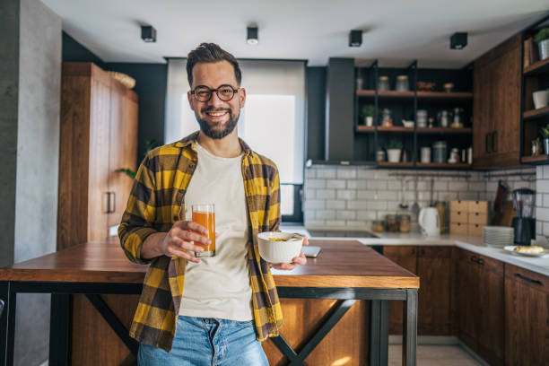 istock Handsome man in glasses standing in kitchen holding bowl of cereal and juice 2193996708