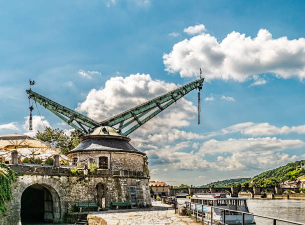 A picturesque riverfront scene featuring a historic crane, a moored boat, and a serene backdrop of a bridge against a beautiful blue sky with scattered clouds.
