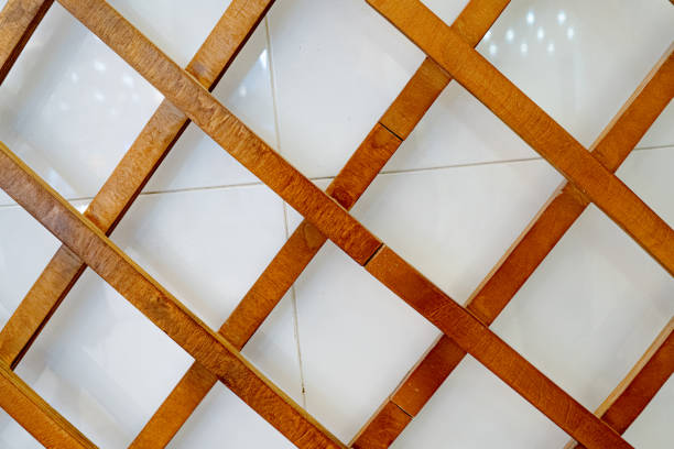 A close-up view of a wooden grid arranged diagonally against a smooth white tiled background. The natural wooden texture and intersecting angles create a visually appealing geometric pattern.