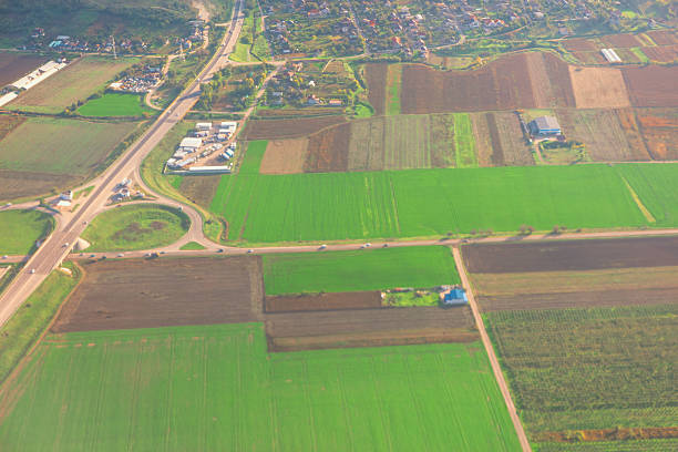 Aerial view of rural area, mix of agricultural fields, roads, small settlement, divided plots, varying crops, roundabout connecting main roads, highlights land use and rural infrastructure