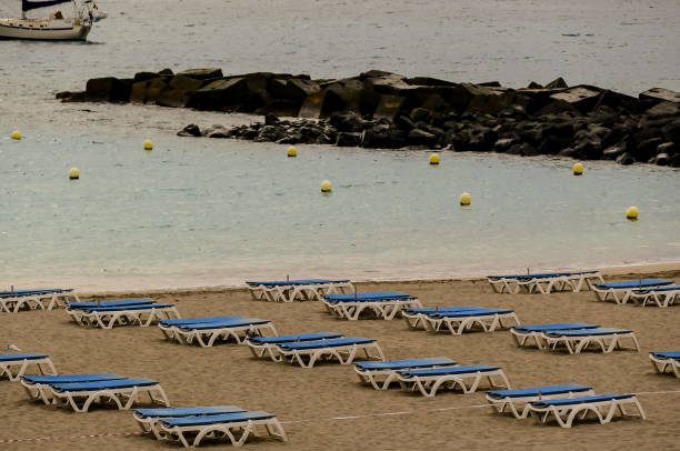 A beach with a large rock wall and many blue lounge chairs. The chairs are empty and the beach is calm