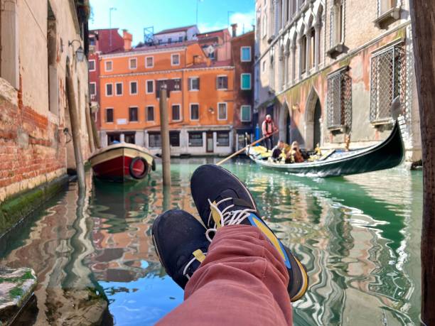 POV Point of view of a tourist is relaxing by a canal in Venice, Italy, with their feet dangling over the water, while a gondola carrying tourists passes by on a sunny day