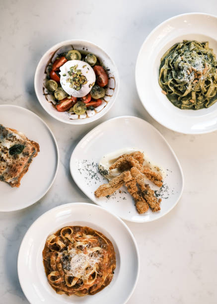 A beautifully arranged selection of traditional Italian dishes displayed on a white marble table. The spread includes Tagliatelle al Ragù alla Bolognese with parmesan, fresh Burrata salad with cherry tomatoes, crispy Trippa Fritta, Melanzane Alla Parmigiana, and creamy Pesto Tagliatelle. Capturing the essence of Mediterranean gastronomy, this image highlights the diversity of Italian cuisine.