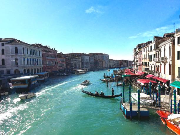 Gondolas and boats are navigating the Grand Canal in Venice, Italy, on a sunny day with colorful buildings lining the waterway and outdoor restaurants bustling with activity