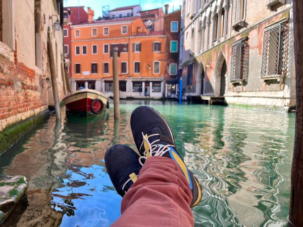 POV Point of view of tourist relaxing with feet dipped in a typical Venetian canal on a sunny day, surrounded by colorful houses and boats, capturing the essence of Italian culture and leisure