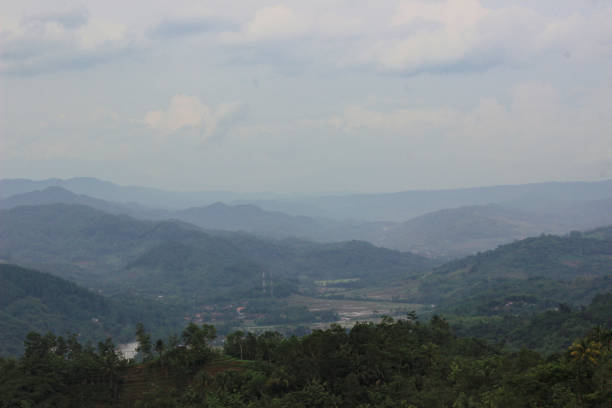 Serene hilly terrain with lush greenery under a cloudy blue sky. The landscape includes distant mountains, forests, and valleys, ideal for nature and outdoor enthusiasts, exuding tranquility and natural beauty.