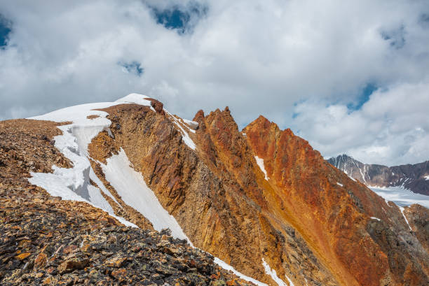 istock Snow-capped mountain ridge with cornice and red sharp rocks on top in sunlight. Snow cap and shiny pointy peak of gold color. Snow dome and colorful rocky peaked top. Sunny cloudy high mountains view. 2194039321