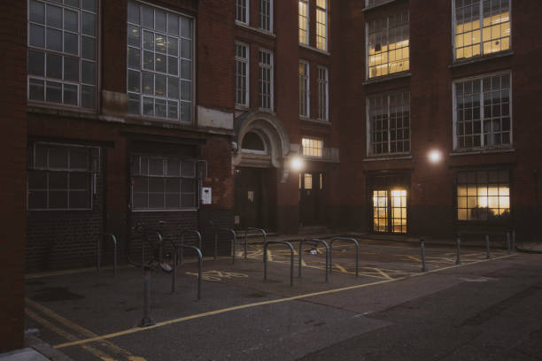 A dimly lit courtyard surrounded by an old brick building with large windows glowing from interior lights at dusk. The scene captures a foggy moody and atmospheric urban environment, with empty bicycle racks and subtle shadows adding to the quiet, contemplative setting. The muted tones and soft lighting evoke a sense of solitude and serenity in London, England, UK
