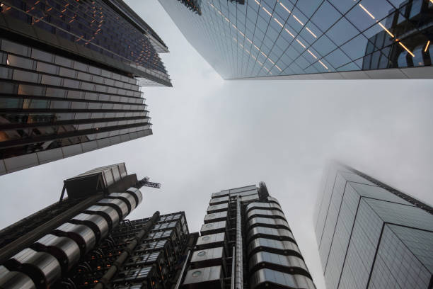 A striking low-angle perspective of London's towering skyscrapers reaching into an overcast sky. The reflective glass facades and industrial-style structures create a futuristic and dynamic cityscape. The mist partially obscures the tops of the high-rises, adding an atmospheric and moody effect. The composition showcases the architectural contrast between sleek modernity and raw industrial design in London, England, UK