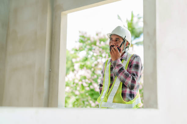 istock Engineer communicating through walkie-talkie near open window of construction site wearing white hard hat and reflective vest. Supervising building progress with scenic background view. 2194042166