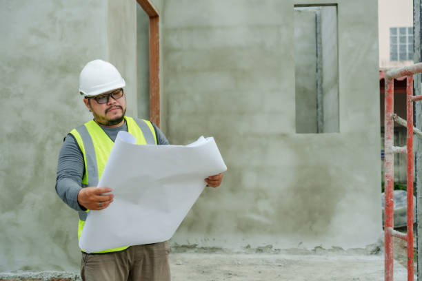 istock Worker holding large blueprint while wearing white safety helmet and bright yellow reflective vest at outdoor construction site with scaffolding and concrete walls, inspection and careful planning. 2194043524