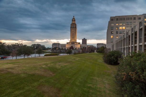 The Louisiana State Capitol building illuminated under a dramatic pre-dawn sky in Baton Rouge, featuring the tranquil surroundings of Capitol Lake and the adjacent historic district.