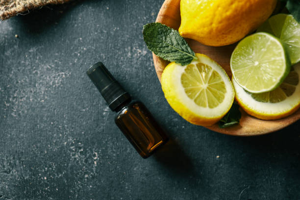 A dark amber essential oil bottle placed on a rustic dark surface with fresh lemons, limes, and mint leaves in the background. A natural and aromatic scene perfect for wellness and organic themes