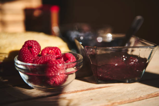 Close-up of fresh raspberries and jam served in bowls on a wooden table. Beautifully captured in natural sunlight, this arrangement is perfect for illustrating fresh ingredients and delightful meals.