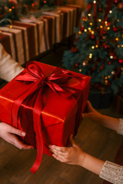 istock Hands exchanging a festive gift box with a red ribbon under a beautifully decorated Christmas tree. Cozy holiday setting with candles, miniature houses, and a warm atmosphere 2194048339