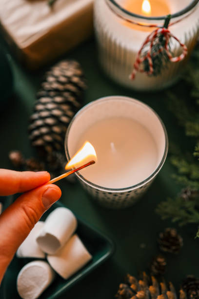 istock A hand lighting a festive candle with a matchstick, surrounded by pinecones, marshmallows, and evergreen branches, creating a warm and cozy Christmas atmosphere 2194049120