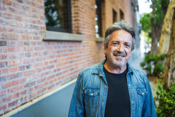 Happy older man standing outdoors by a brick wall on a sunny day. Looking at the camera