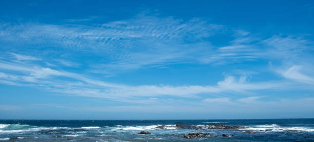 Coastal scene, gentle ocean waves breaking against rocky shore under a vibrant blue sky with scattered clouds.