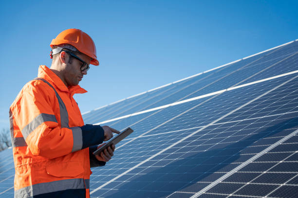 istock Industrial worker in orange safety gear reviews solar panel system with device on bright sunny day. 2194055255