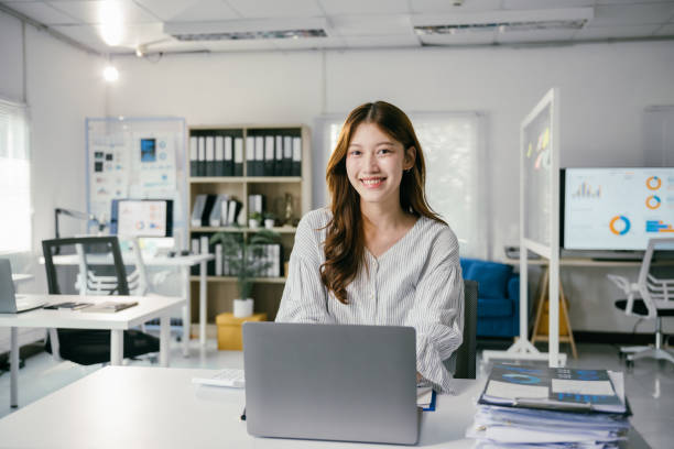 istock Young asian businesswoman smiling while working on a laptop in a modern office, sitting confidently at her desk and engaging with the camera, exuding professionalism and positivity 2194058837