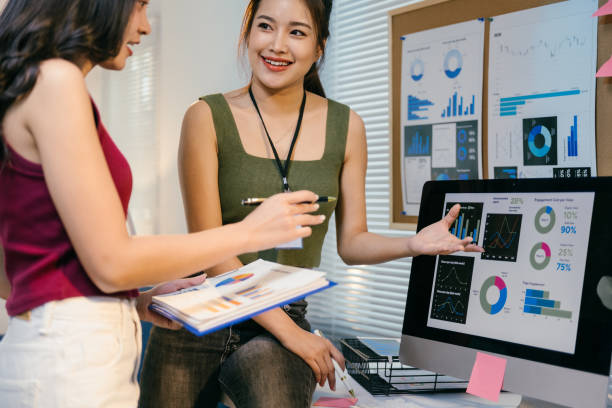 istock Two young businesswomen analyzing marketing data on a computer screen, pointing at charts and graphs while holding a folder of reports in a modern office 2194064440