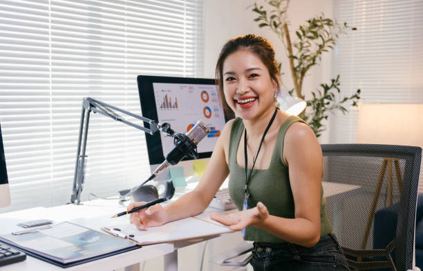 istock Young asian businesswoman smiling while recording a podcast in her home office, using a professional microphone and jotting down notes at her desk, enjoying the creative process 2194064576