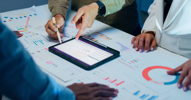 istock Close-up group of business working late, analyzing financial reports and charts on digital tablet surrounded by documents and graphs during strategic meeting at office night. Finance meeting. 2194068228