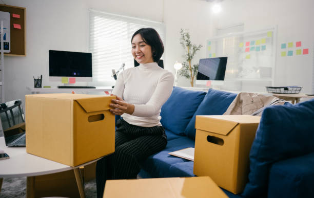 istock Young asian businesswoman sitting on a blue sofa, smiling while packing cardboard boxes for a move to a new office, embodying the excitement of starting a new chapter in her career 2194074683