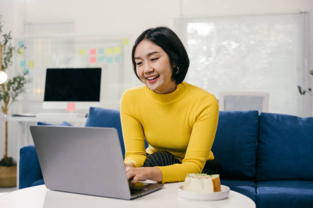 istock Happy young asian woman working remotely from home, sitting on a comfortable sofa and typing on her laptop, enjoying a sandwich during her break 2194074714