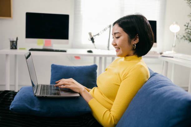 istock Happy young asian woman working from home, telecommuting or studying online using laptop computer while sitting comfortably on a blue couch in her living room 2194074767