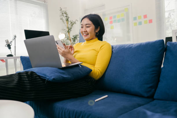 istock Cheerful young asian freelancer woman using laptop computer and smartphone while relaxing on comfortable sofa, enjoying work from home and modern lifestyle 2194074785